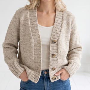May include: A beige button-down cardigan sweater with a ribbed collar and cuffs. The sweater has a textured knit pattern and is buttoned with four brown buttons. The model is wearing a white tank top and blue jeans.