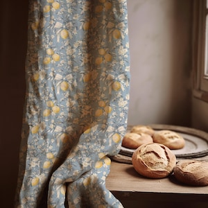 May include: A light blue fabric curtain featuring a repeating pattern of yellow lemons, white floral sprigs, and tied bows. The textile hangs vertically, displaying a detailed botanical print with soft color contrasts and a textured appearance.