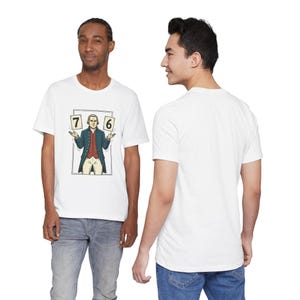 Op de afbeelding: Wit T-shirt met een cartoon illustratie van een man die kaarten vasthoudt met de nummers 7 en 6. De man in de illustratie draagt een blauwe jas, een rood vest en een witte broek. Het T-shirt wordt door een persoon gedragen.