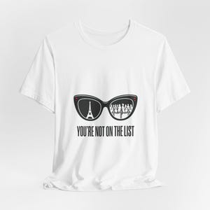 Op de afbeelding: Wit t-shirt met een zwarte afbeelding van een zonnebril, de Eiffeltoren en een menigte. De tekst "YOU'RE NOT ON THE LIST" staat eronder in het zwart.