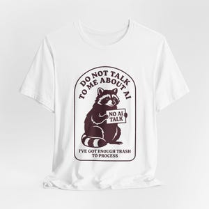 Op de afbeelding: Wit t-shirt met een wasbeer-afbeelding. De wasbeer houdt een bord vast met de tekst "NO AI TALK". Het shirt heeft ook de tekst "DO NOT TALK TO ME ABOUT AI" en "I'VE GOT ENOUGH TRASH TO PROCESS."