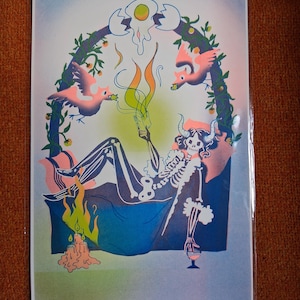 May include: A rectangular art print featuring a horned skeleton reclining on a blue surface, holding a book with green flames rising from it. The scene includes two pink birds, a cracked egg at the top, and three lit candles near a wine glass.