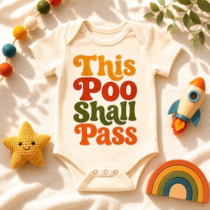 May include: Cream-colored baby onesie with the text "This Poo Shall Pass" in orange, red, and green. The onesie is surrounded by a star-shaped toy, a wooden rocket, a rainbow, and a garland of colorful felt balls.