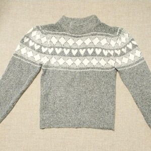 May include: A gray long-sleeve sweater featuring a mock neck and a white patterned yoke. The yoke displays a repeating design of diamonds and hearts across the chest and shoulders. The sweater has a textured knit finish and ribbed cuffs.