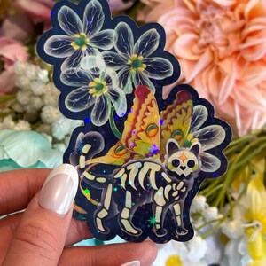 May include: A holographic sticker featuring a skeletal cat with a skull-shaped face and large, colorful moth wings. The cat is positioned beneath three translucent, glowing white flowers against a dark blue background with scattered star accents.