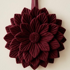May include: A deep burgundy velvet flower wall hanging featuring layered, pointed petals arranged in a circular pattern. The center of the flower has a small, textured pom-pom detail. A matching burgundy ribbon is attached to the top for hanging.