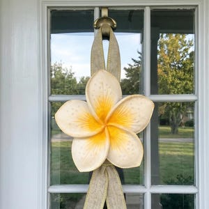 May include: A large, cream-colored plumeria flower wreath with a yellow center hangs on a white door. The flower is attached to a tan ribbon, creating a welcoming floral decoration. The door has glass panes, reflecting the outdoor scenery.