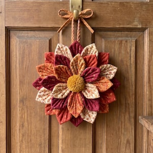 May include: A fabric flower wreath in autumnal colours of orange, red, and cream, with a yellow centre, hanging on a wooden door. The door has a brass handle and is made of wooden panels.
