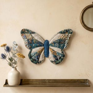 May include: A decorative butterfly wall hanging with a patchwork design in shades of blue, green, and cream. The butterfly is mounted on a beige wall, with a small round mirror and a shelf with a vase of flowers.
