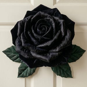 May include: A large, plush, decorative rose-shaped pillow crafted from black velvet fabric. The flower features layered, soft petals arranged in a circular bloom pattern, accented by several dark green, leaf-shaped fabric pieces at the base.