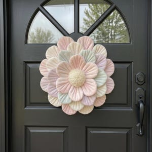May include: A decorative fabric flower wreath in pastel colours hangs on a black front door. The wreath is circular, with layers of petals in shades of pink, yellow, and blue. The door has a window at the top and a black handle.