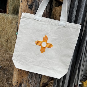 Peut inclure: Un sac fourre-tout en toile couleur crème avec un symbole solaire Zia orange brodé au centre sur le devant. Le sac possède deux anses en tissu assorti fixées au bord supérieur et une petite étiquette verte sur la couture latérale gauche.