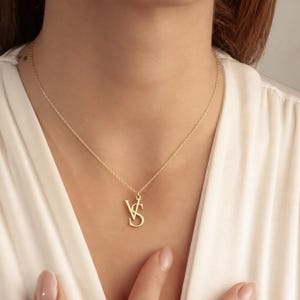 May include: A gold-toned necklace featuring a delicate chain and a pendant consisting of the capital letters V and S intertwined. The pendant has a polished finish and hangs centrally from the chain, resting against a white fabric garment.