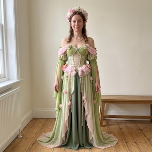 Può includere: Un abito lungo fino ai piedi con spalle scoperte in tonalità verde salvia e oliva con accenti in chiffon rosa pallido. Il corpetto presenta un'allacciatura frontale, mentre la gonna e le maniche sono decorate con fiori in tessuto e perline.