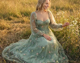 Könnte beinhalten: Ein salbeigrünes, bodenlanges Kleid mit herzförmigem Ausschnitt, langen, transparenten Puffärmeln und einem figurbetonten Oberteil. Das Kleid ist mit zarten, mehrfarbigen Blumenstickereien auf Rock und Ärmeln verziert, die eine weiche Textur erzeugen.
