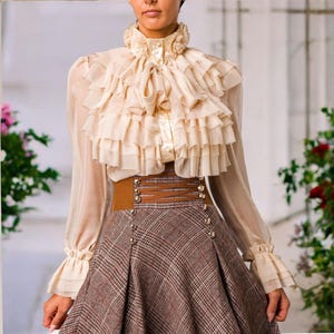 May include: A cream-colored long-sleeve blouse featuring a high ruffled collar, layered ruffles across the chest, and flared cuffs, paired with a brown plaid skirt that has a wide corset-style waistband with brown lacing and silver buttons.