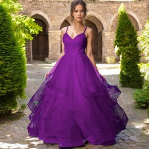 May include: A floor-length, vibrant purple gown featuring a fitted bodice with a V-neckline, thin spaghetti straps, and a voluminous, multi-layered tulle skirt that cascades down to the floor in soft, flowing ruffles.
