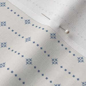 May include: Close-up of white fabric with a repeating pattern of blue floral and geometric designs. Two pins are inserted into the fabric, holding it together. The fabric has a subtle texture and a clean, crisp appearance.