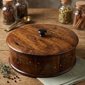 Könnte beinhalten: Runde Gewürzbox aus dunklem Holz mit dekorativen goldenen Intarsien. Der Deckel hat einen schwarzen Knopf. Gewürzgläser und ein kleiner Messinglöffel sind im Hintergrund zu sehen, was auf eine Küche oder ein kulinarisches Thema hindeutet.