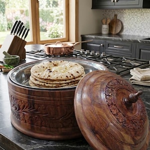 May include: A wooden roti server with a stack of flatbread inside. The server has a carved floral design and a matching lid. The server is made of dark brown wood and sits on a black countertop. A copper pan is visible in the background.