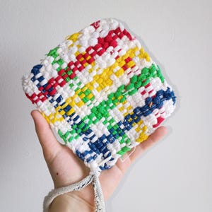 May include: A colorful, woven potholder in a square shape. The potholder features a pattern of red, yellow, green, blue, and white threads. A loop is attached for hanging. The potholder is held in a hand against a plain white background.