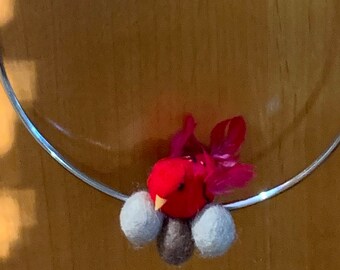 Pode incluir: Um aro de metal prateado com um pássaro de feltro vermelho e três bolas de feltro branco penduradas nele. O aro está suspenso por um cordão branco.