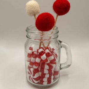 Könnte beinhalten: Ein klares Glas mit Griff, gefüllt mit rot-weiß gestreiften Bonbons. Drei Filzkugel-Lollis, zwei rote und einer cremefarbener, befinden sich im Glas. Die Lollis sind auf Holzstäbchen.