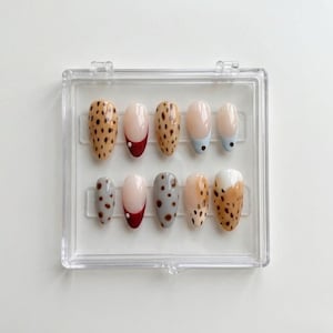 May include: A set of ten press-on nails arranged in a clear plastic storage case. The nails feature various designs including brown leopard print, solid nude with red tips, solid grey with brown spots, and a white and brown abstract pattern.