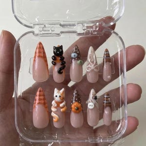 May include: A set of ten stiletto-shaped press-on nails in a clear plastic case. The designs include orange plaid tips, 3D cat figures, brown polka dots, white swirl patterns, a small orange pumpkin, a white button accent, and brown striped tips.