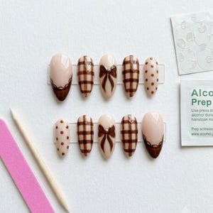 May include: A set of ten almond-shaped press-on nails in a nude and brown color palette. The designs include brown plaid patterns, brown bows, brown polka dots, and brown scalloped tips. Included are a pink nail file, a wooden stick, and adhesive tabs.