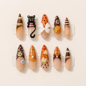May include: A set of ten stiletto-shaped press-on nails featuring Halloween-themed designs. The nails include motifs such as a black cat, a white bow, a pumpkin, buttons, plaid patterns, polka dots, and stripes in orange, brown, and cream colors.