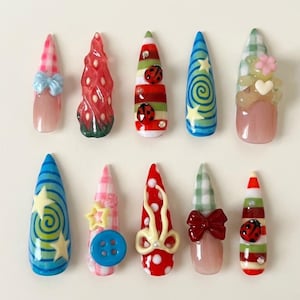 May include: A set of ten press-on nails featuring various designs including a pink gingham pattern with a blue bow, a red strawberry texture, green and red stripes with ladybugs, blue swirls with stars, and a bear with a heart and flower.