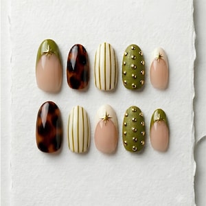 May include: A collection of ten artificial nails with various designs. The designs include olive green with gold star accents, tortoiseshell, vertical stripes, and olive green with gold studs. Some nails have a nude base with a white or olive green tip.