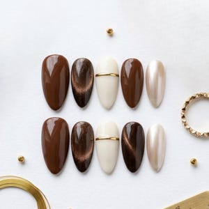 May include: A set of ten oval-shaped false nails in various shades of brown, beige, and gold. The nails are arranged in two rows of five, with a mix of solid colors, cat-eye effects, and a gold stripe accent. A gold ring and small gold beads are also present.