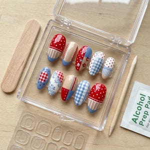 May include: A set of ten press-on nails in a clear plastic case, featuring various patterns including red and white polka dots, blue and white gingham, and floral designs. The kit includes a wooden cuticle stick and an alcohol prep pad.