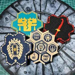 Op de afbeelding: Een verzameling speltoken met verschillende ontwerpen en kleuren. Eén token is turquoise met een gouden drakenontwerp, een ander is zwart met een rood zwaard, en andere bevatten tekst zoals "FIGHT FIRST" en "FIGHT LAST". De tokens zijn gerangschikt op een grijs, getextureerd oppervlak.