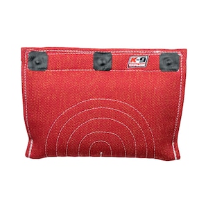 May include: A rectangular red dog bite training pillow featuring a textured fabric surface with white reinforced stitching. The top edge includes three black circular fasteners and a white label with the black and red text K-9 REDLINE.