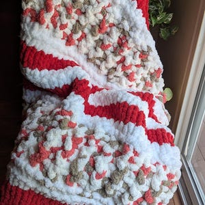 May include: A chunky knit blanket featuring a thick, textured pattern with alternating stripes of solid red, bright white, and a variegated blend of white, tan, and coral yarn. The soft, plush material is crafted with large, visible loops throughout.