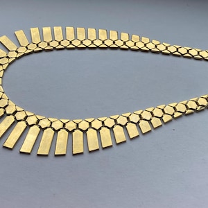 Op de afbeelding: Een goudkleurige metalen ketting met een reeks verbonden zeshoekige schakels die overgaan in een franje van platte, rechthoekige, verticale staven aan de voorzijde, wat een gestructureerd, stralend kraagontwerp met een metallic afwerking creëert.