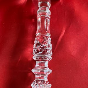 Op de afbeelding: Een heldere, kleurloze glazen hamer met ingewikkelde geslepen patronen. De kop van de hamer heeft een ruitvormig ontwerp, terwijl het handvat een reeks decoratieve, gefacetteerde secties vertoont die toelopen naar een puntige, geribbelde basis.