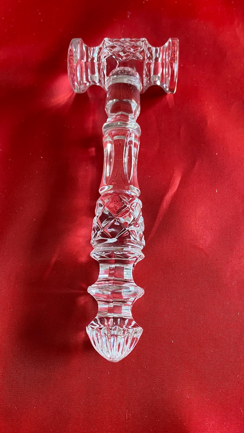 Op de afbeelding: Een heldere, kleurloze glazen hamer met ingewikkelde geslepen patronen. De kop van de hamer heeft een ruitvormig ontwerp, terwijl het handvat een reeks decoratieve, gefacetteerde secties vertoont die toelopen naar een puntige, geribbelde basis.
