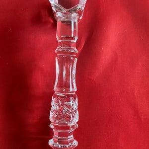 Op de afbeelding: Een heldere kandelaar van geslepen glas met een gefacetteerde, zeshoekige opening aan de bovenkant en een decoratieve, uit meerdere delen bestaande steel met ingewikkelde kruisarceringen en verticale patronen. De voet is afgerond en getextureerd, met het woord Waterford erin gegraveerd.