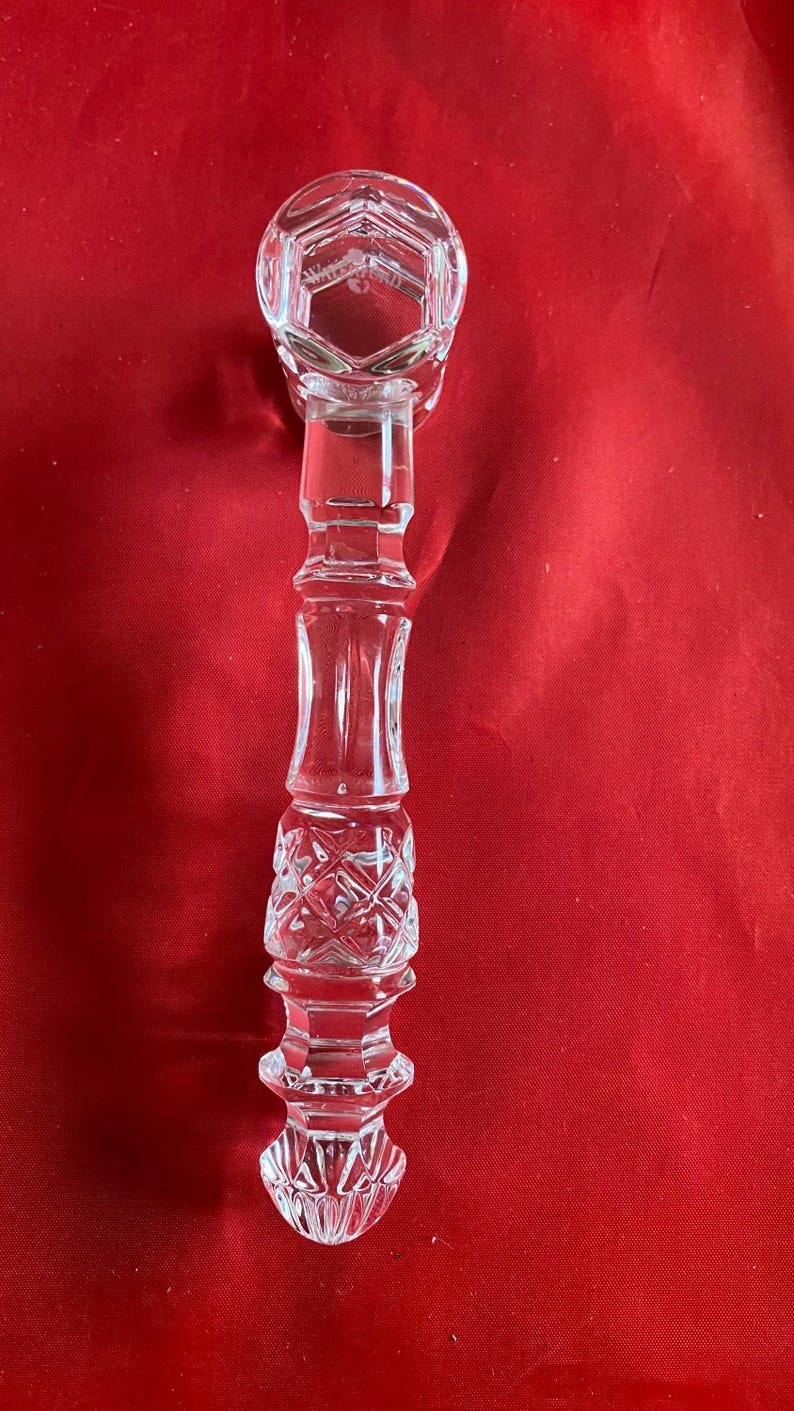 Op de afbeelding: Een heldere kandelaar van geslepen glas met een gefacetteerde, zeshoekige opening aan de bovenkant en een decoratieve, uit meerdere delen bestaande steel met ingewikkelde kruisarceringen en verticale patronen. De voet is afgerond en getextureerd, met het woord Waterford erin gegraveerd.