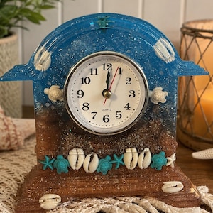 May include: A resin mantel clock featuring a gradient design transitioning from a sandy brown base to a glittery blue top. The clock face is white with black numerals and hands. It is decorated with embedded white seashells and small turquoise sea turtles.