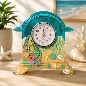May include: A teal resin mantel clock with an arched top and a round white face featuring black Arabic numerals and red and black hands. The base contains embedded sand, a sculpted mermaid figure, small starfish, a turtle, and seashells.