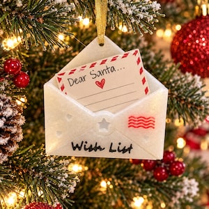 May include: A white, envelope-shaped Christmas ornament with a gold ribbon hanger. The front features the text "Wish List" in black, a red stamp graphic, and a star cutout. A letter inside reads "Dear Santa..." with a red heart and lines.