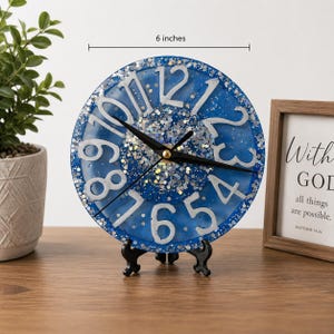 May include: A circular blue resin clock with silver glitter accents and raised white numbers. The clock face features black hour, minute, and second hands. A measurement indicator above the clock shows a diameter of 6 inches. It rests on a black stand.