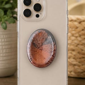 May include: A clear smartphone case featuring a circular phone grip attached to the back. The grip has a smooth, glossy surface with a swirling pattern of metallic copper, bronze, and dark gray tones, creating a marbled effect across the center.