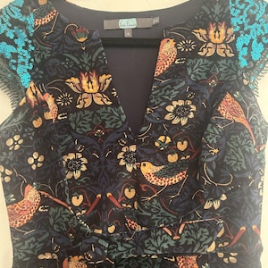 K&ouml;nnte beinhalten: Ein marineblaues Kleid mit V-Ausschnitt und kurzen &Auml;rmeln, mit einem Blumen- und Vogelmuster in Orange-, Gr&uuml;n- und Cremet&ouml;nen. Die &Auml;rmel sind mit t&uuml;rkisfarbenen Pailletten verziert. Das Kleid h&auml;ngt an einem schwarzen Kleiderb&uuml;gel.