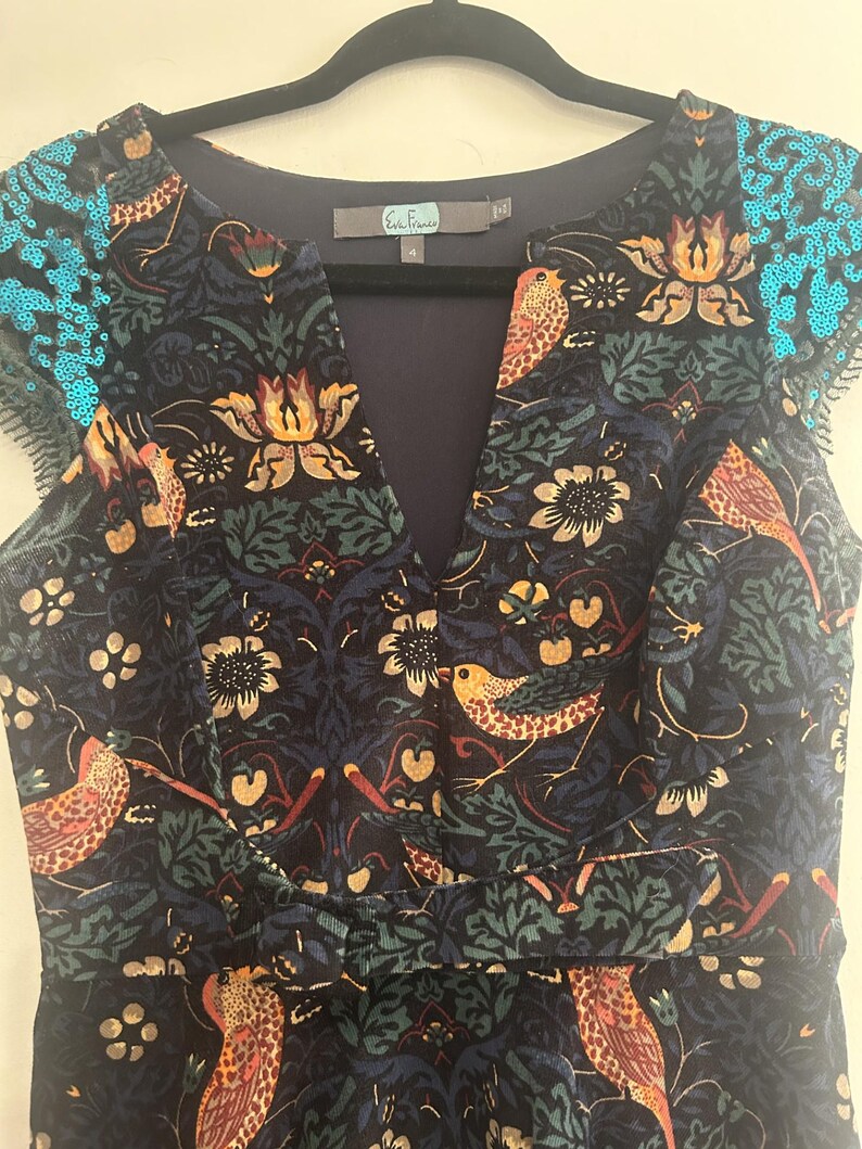 K&ouml;nnte beinhalten: Ein marineblaues Kleid mit V-Ausschnitt und kurzen &Auml;rmeln, mit einem Blumen- und Vogelmuster in Orange-, Gr&uuml;n- und Cremet&ouml;nen. Die &Auml;rmel sind mit t&uuml;rkisfarbenen Pailletten verziert. Das Kleid h&auml;ngt an einem schwarzen Kleiderb&uuml;gel.